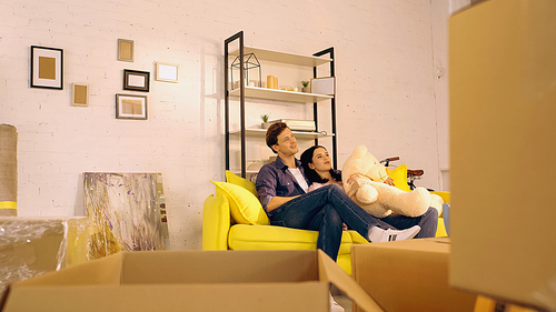 cheerful couple chilling on sofa with teddy bear near boxes in new home