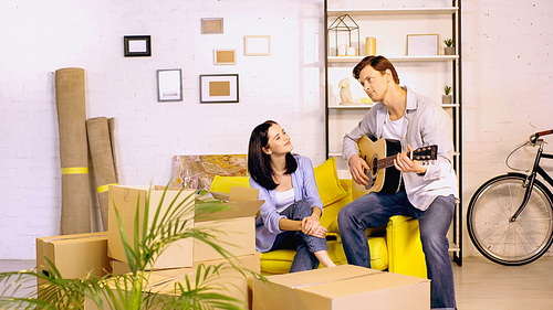 man playing acoustic guitar near happy girlfriend sitting on couch in new home