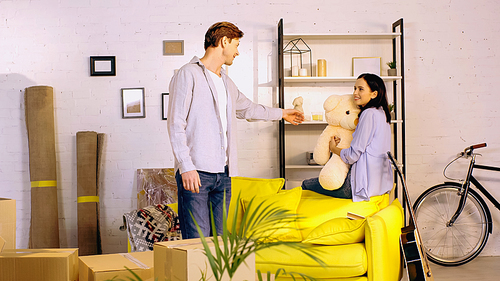 happy man reaching woman with teddy bear in living room