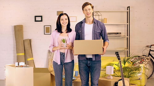 Cheerful couple with box and plant in new home