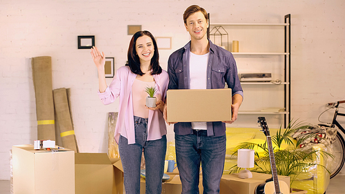 happy woman waving hand and holding plant near man with box in new home