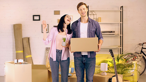 Cheerful woman waving hand and holding plant near man with box in new home