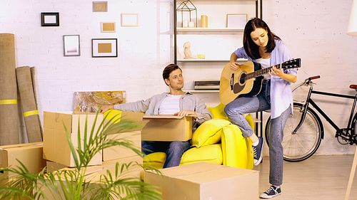 young woman playing guitar near boyfriend with box in new home