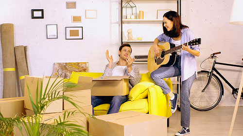 young woman playing guitar near boyfriend applauding in new home