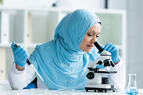 Arabian scientist in hijab looking through microscope and holding electronic pipette