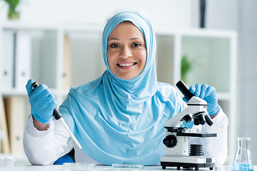 Smiling arabian scientist holding pipette and microscope while working in lab