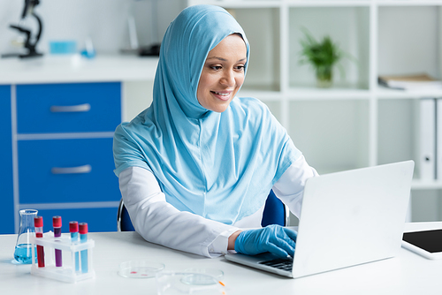 Positive arabian scientist using laptop near test tubes, petri dishes and digital tablet