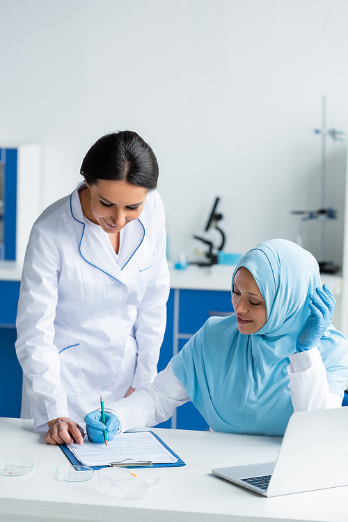 Smiling arabian scientist writing on clipboard near laptop, colleague and petri dishes