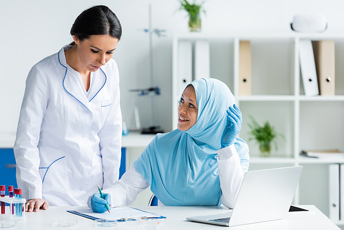 Cheerful muslim scientist in latex gloves writing on clipboard and looking at colleague near laptop in laboratory