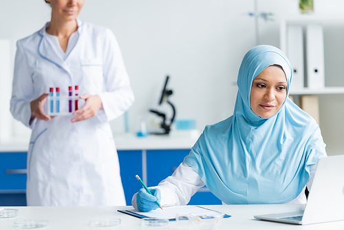 Muslim scientist writing on clipboard and looking at laptop near blurred colleague with test tubes