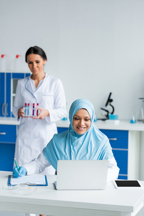 Smiling muslim scientist using laptop and writing on clipboard near blurred colleague