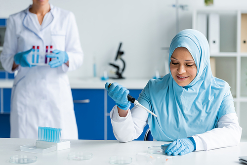 Smiling arabian scientist holding electronic pipette near petri dishes and colleague