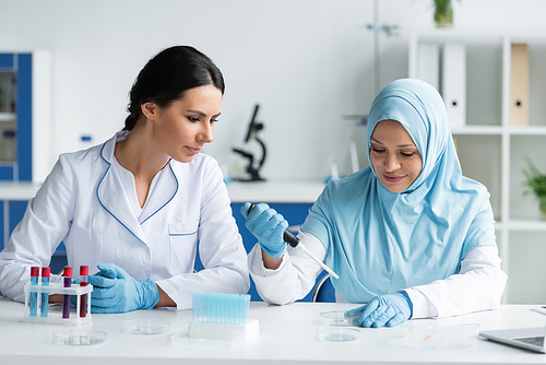 Multiethnic scientists in medical masks working with petri dishes and electronic pipette near blurred laptop