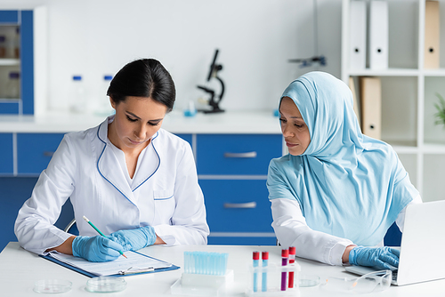 Scientist in latex gloves writing on clipboard near arabian colleague using laptop in laboratory