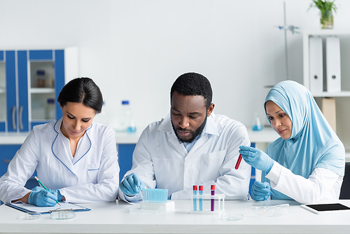 Interracial scientists working with test tubes near colleague writing on clipboard in lab