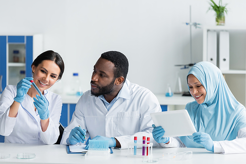 Smiling scientist holding test tube near interracial colleagues with clipboard and digital tablet