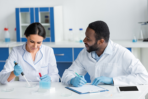 Smiling african american scientist in latex gloves writing on clipboard and looking at colleague with test tube