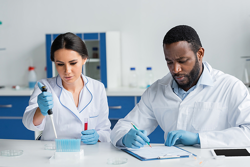 African american scientist writing on clipboard near colleague with pipette and test tubes