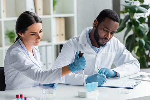 Scientist holding electronic pipette near test tubes and blurred african american colleague writing on clipboard