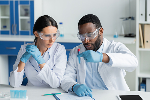 Interracial scientists in safety goggles holding test tube near clipboard and digital tablet