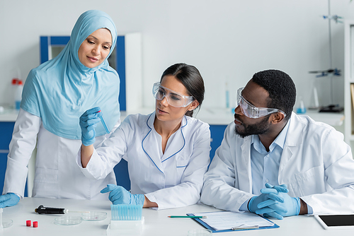 Scientist holding test tube near interracial colleagues in safety goggles, electronic pipette and digital tablet