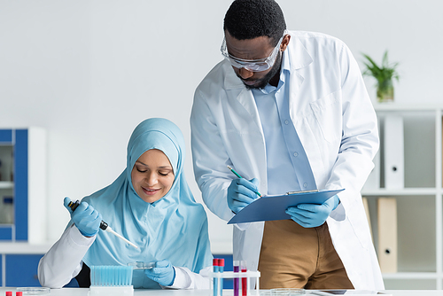 African american scientist in safety goggles writing on clipboard near smiling arabian colleague with pipette and petri dish