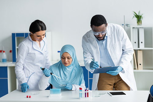 Multiethnic scientists in latex gloves working with clipboard and petri dishes in laboratory