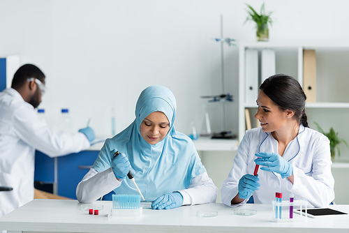 Smiling scientist holding blood sample near arabian colleague with petri dish in laboratory