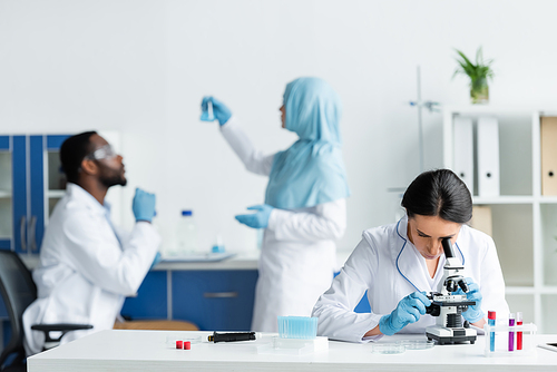 Scientist looking through microscope near blurred interracial colleagues in lab