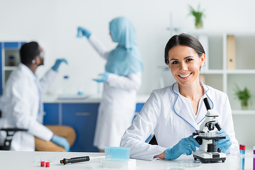 Happy scientist in latex gloves smiling at camera near microscope and test tubes