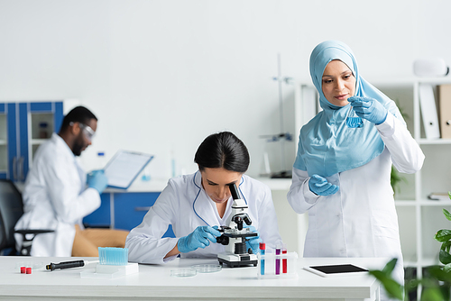 Arabian scientist holding flask with reagent near colleague with microscope