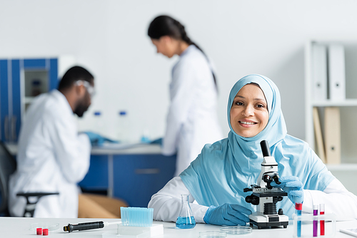 Positive muslim scientist looking at camera near microscope, flask and pipette in lab