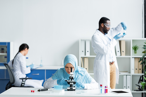 Muslim scientist looking through microscope near digital tablet and interracial colleagues on blurred background