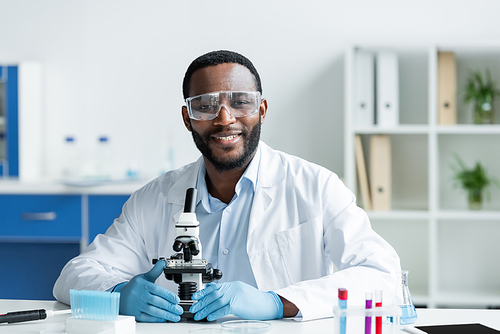 Smiling african american scientist in protective goggles looking at camera near microscope in lab