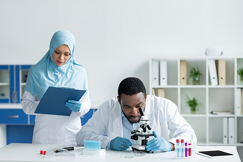 African american scientist looking through microscope near muslim colleague with clipboard