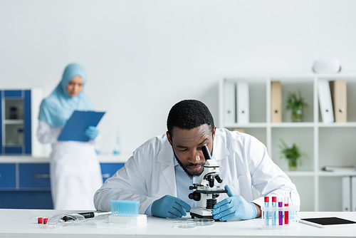 African american scientist in white coat looking through microscope near test tubes and digital tablet