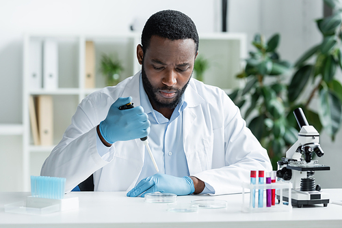 African american scientist in latex gloves holding electronic pipette near petri dish and microscope