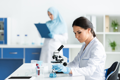 Scientist in medical mask taking flask near microscope and test tubes