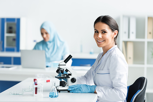 Smiling scientist in latex gloves looking at camera near microscope and equipment