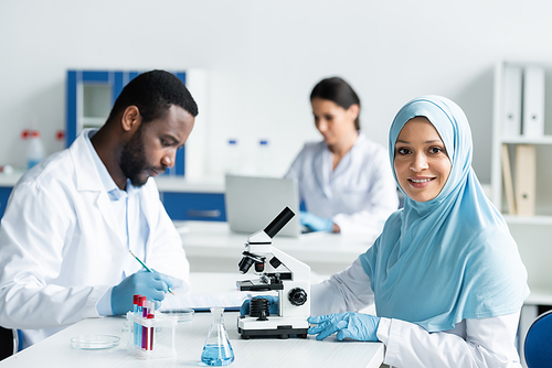 Smiling arabian scientist in hijab looking at camera near microscope and african american colleague in laboratory