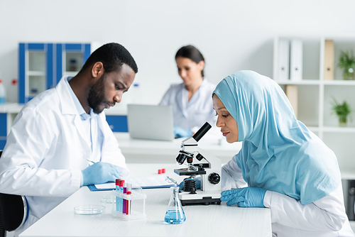 Arabian scientist working with microscope near test tubes and blurred colleagues