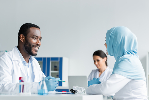 African american scientist smiling at muslim colleague near equipment in laboratory