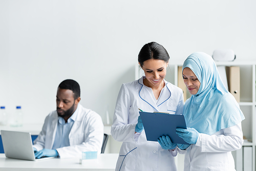 Interracial scientists smiling while holding clipboard near blurred african american colleague with laptop in lab