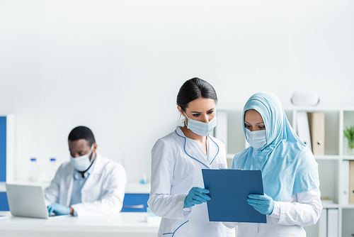 Interracial scientists in medical masks looking at clipboard near african american colleague