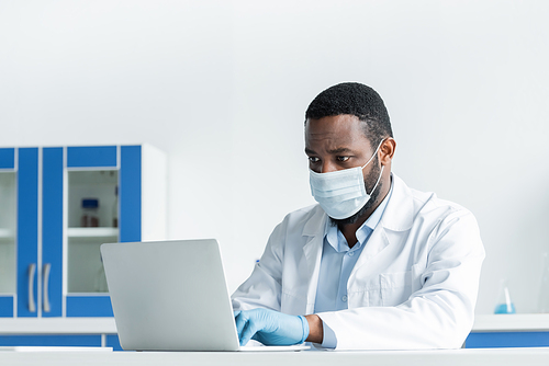 African american scientist in medical mask using laptop in laboratory