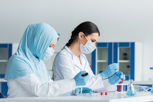 Side view of scientist in protective mask working near arabian colleague with pipette on blurred foreground