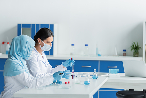 Side view of interracial scientists in protective masks working with medical equipment near laptop in lab