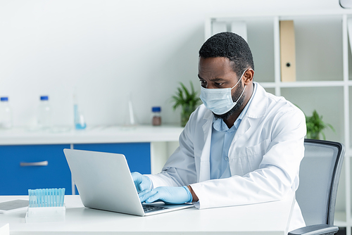 African american scientist in latex gloves and medical mask using laptop near test tubes in lab