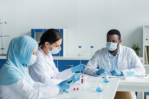 African american scientist with clipboard looking at blurred interracial colleagues with test tubes