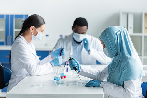 Arabian scientist in latex gloves using electronic pipette and petri dishes near colleagues